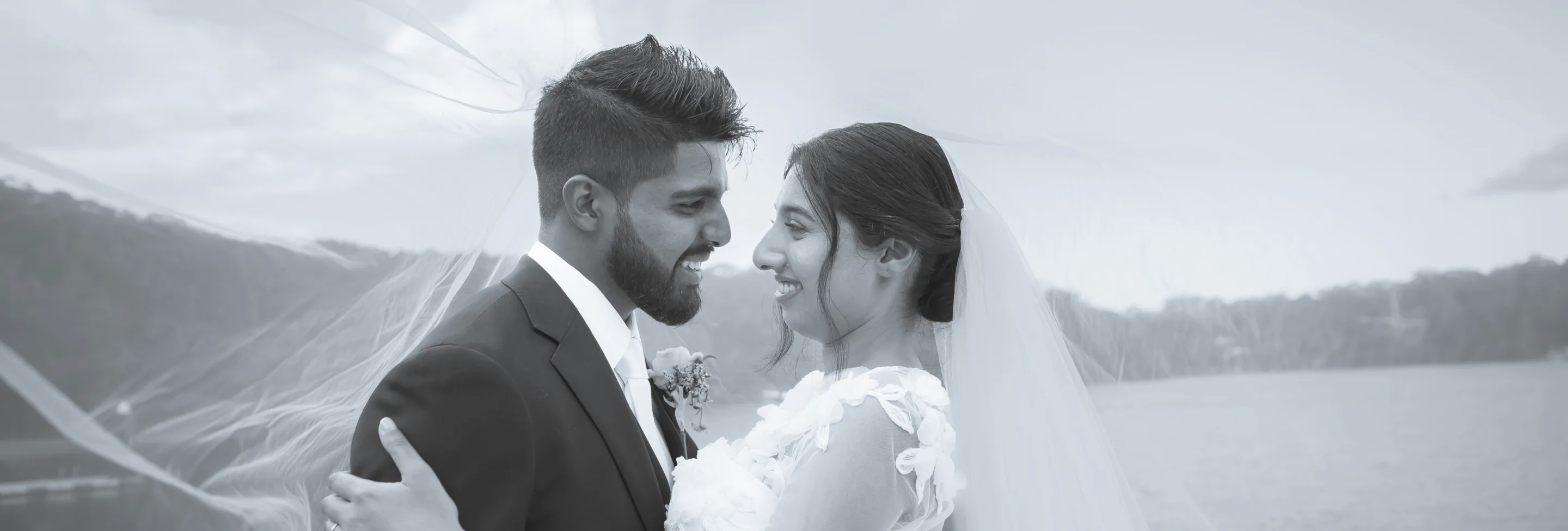 Bride and groom facing each other by the water, veil in the wind