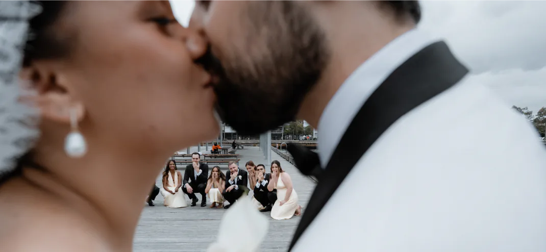 Bride and groom kissing in the foreground with their wedding party watching from a boardwalk